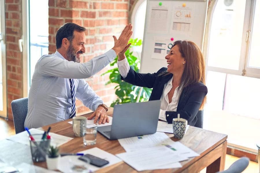 Colleagues high-five over a laptop and papers, highlighting 24/7 Accessibility in their sunlit office workspace.