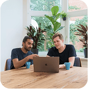 Two men reviewing financial data on an investor website using a laptop in a modern office with plants.