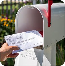 Hand placing official election material and notice mailing into an open white U.S. Mail mailbox.