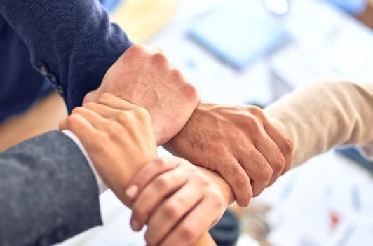 Four team members join hands in a wrist-to-wrist grip representing unity and flexible solutions for business.