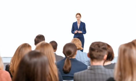Female executive presenting to a professional audience during shareholder meetings in a bright, modern setting.